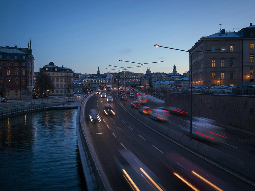 trafik i kvällsljus på trafikled genom Stockholms stad, med flera bostadshus intill vägen