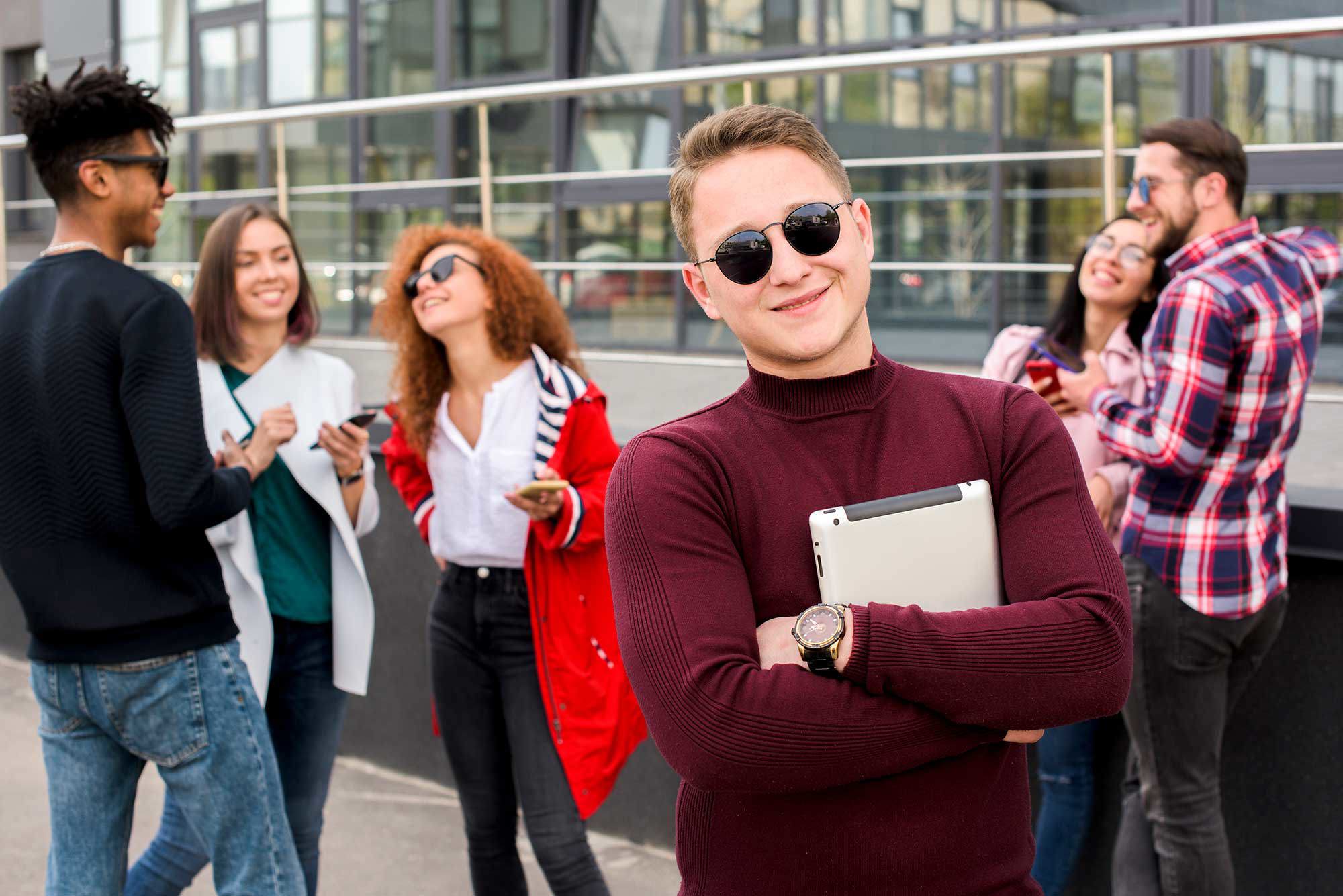 Student som står i solglasögon och tittar in i kameran, i omgiven av andra studenter
